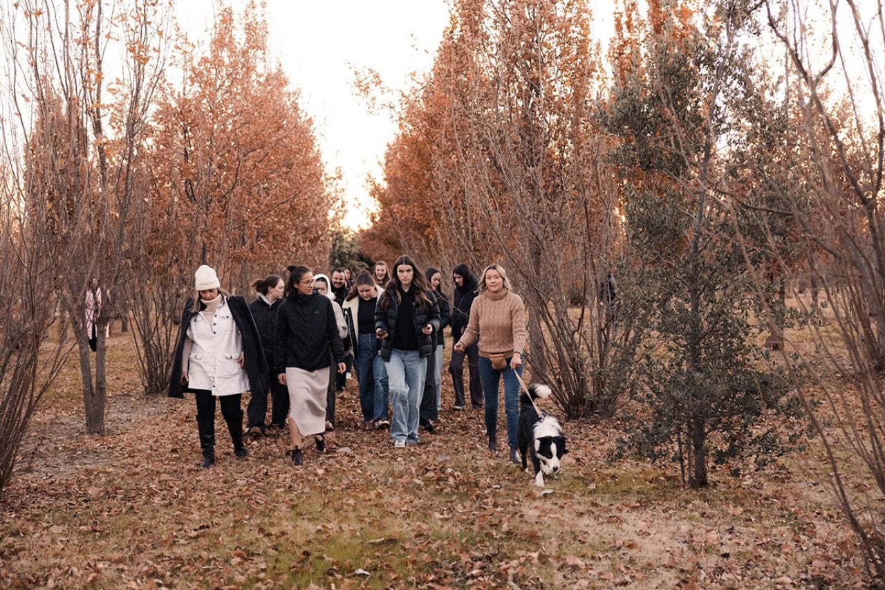 A group of people dressed in fall attire and a dog on a leash walking through trees on a tour of a truffle farm.