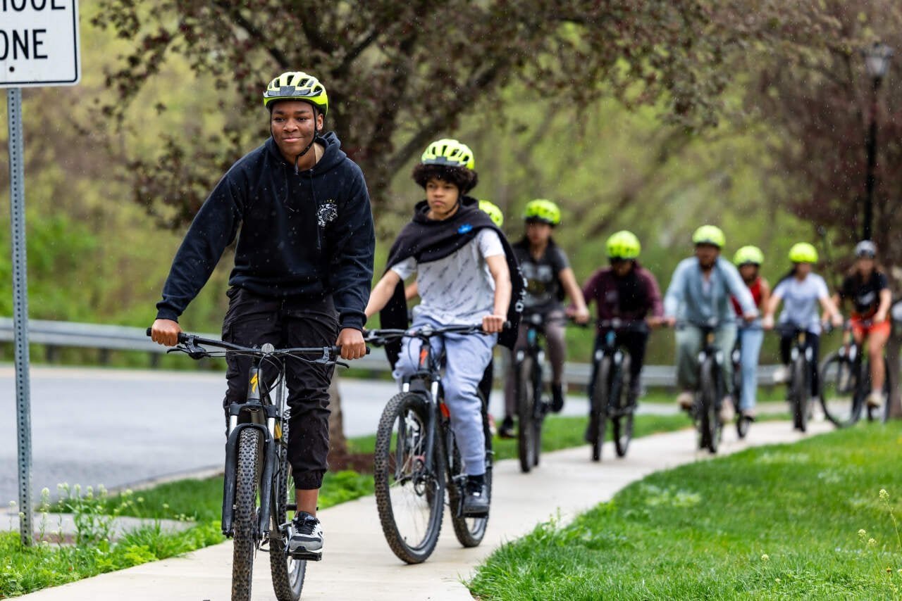 Students on bicycles from Martin Luther King, Jr. Middle School in Germantown, Maryland wearing bright green helmets ride in a line on a sidewalk with grass one one side and trees on the other.