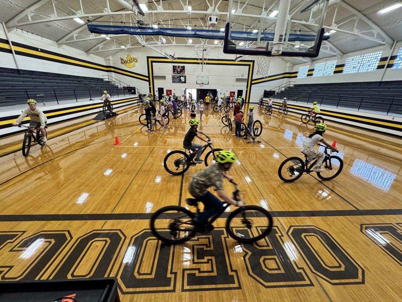 Students riding bikes indoors in a big gym with wooden floors and an arched ceiling in Ashland, Wisconsin.