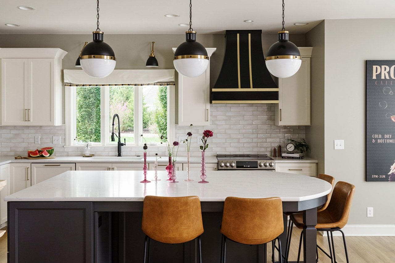 Kitchen with three round hanging light fixtures, a black and gold range hood, white tile and in the foreground a gently rounded kitchen counter stools around an island curved at one end.