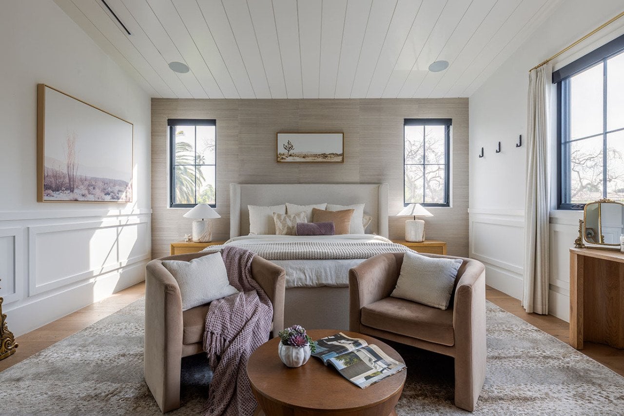A bedroom decorated in warm neutral tones of pale blush, beige and mauve with two velvet chairs in the foreground in front of a bed, featuring an accent wall in a material with a luxurious feel.
