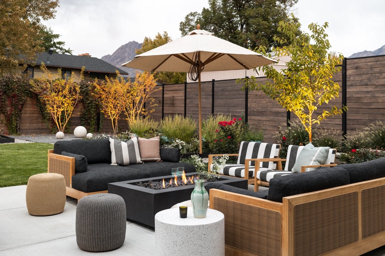 Backyard patio set in neutral black and tan colored furniture arranged around a low gas firepit and trees and horizontal wooden fence in the background.
