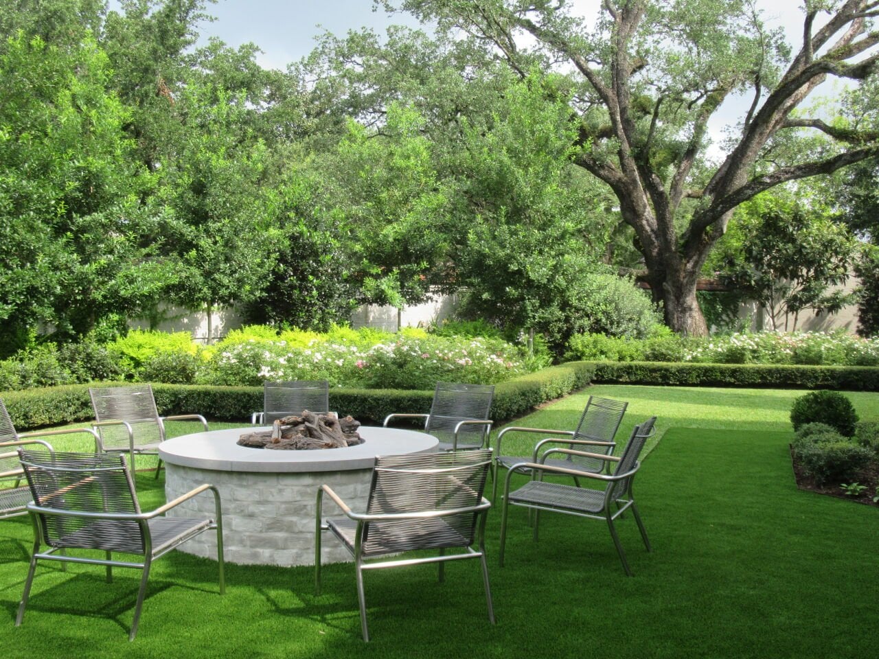 Lush green yard with trees, a low hedge and manicured lawn featuring nine wide metallic chairs around a circular firepit in the foreground.