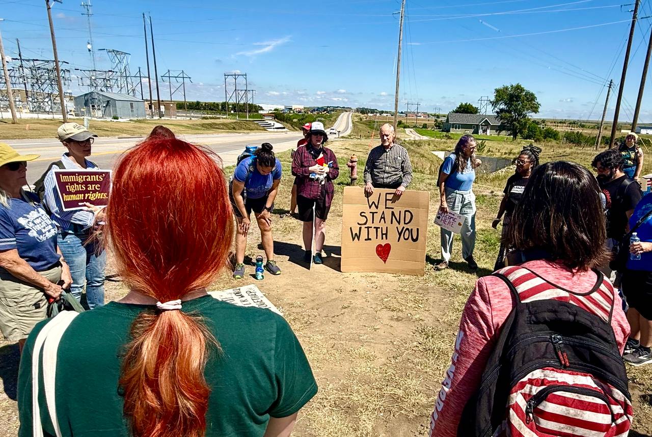 A group traveling from Lincoln to the McCook rally stands in a circle to pray, with some holding signs of support for immigrants. The McCook immigration detention facility is down the highway about a quarter of a mile.
