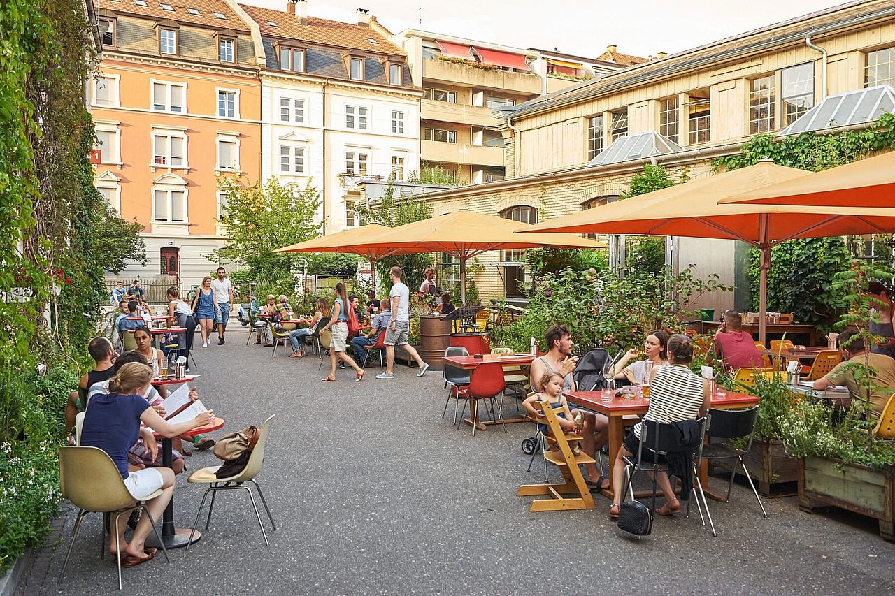 People sitting in chairs and tables outside under large orange umbrellas outside a project designed by Buser.