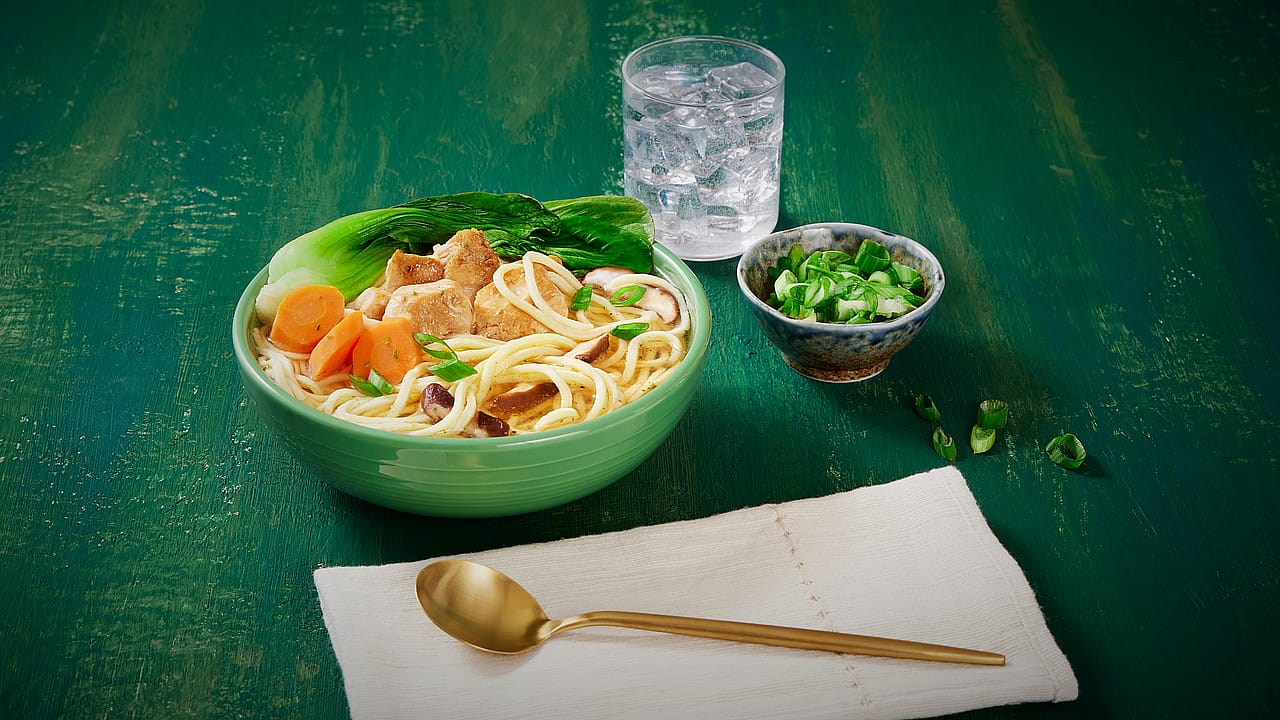 A chicken noodle soup topped with bok choy, carrots, mushrooms with minced green onions on the side.