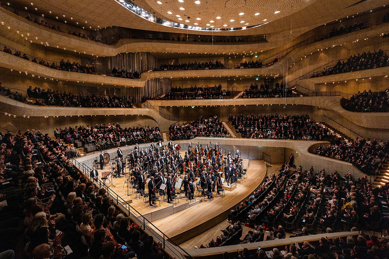 Interior view of the Elbphilharmonie concert hall with an orchestra standing on a stage and audience seated in theater in the round.