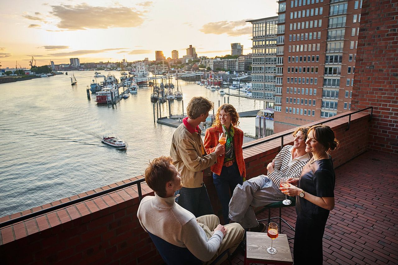 A group of five people enjoy drinks on an outdoor patio overlooking a river near the Elbphilharmonie.