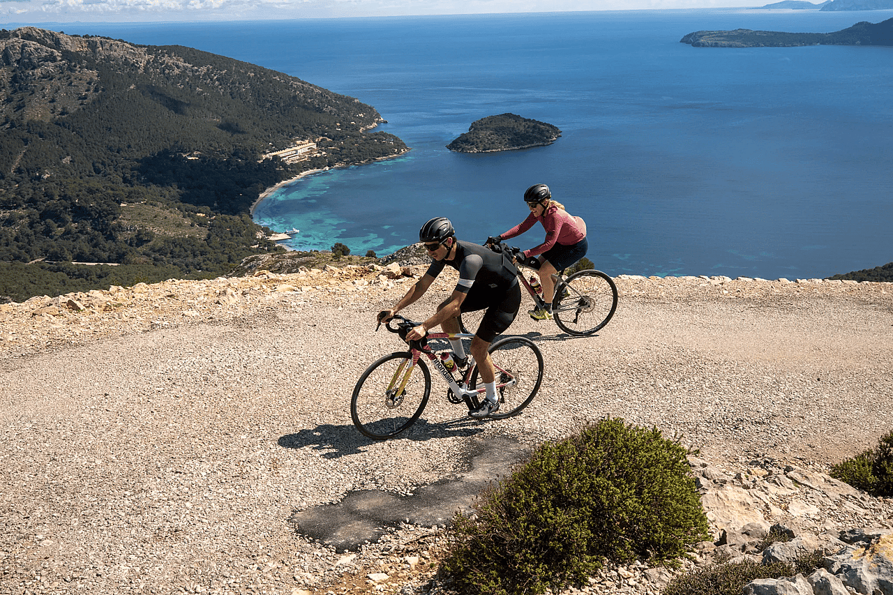 Two people biking on a wide gravel path near the ocean in Mallorca.