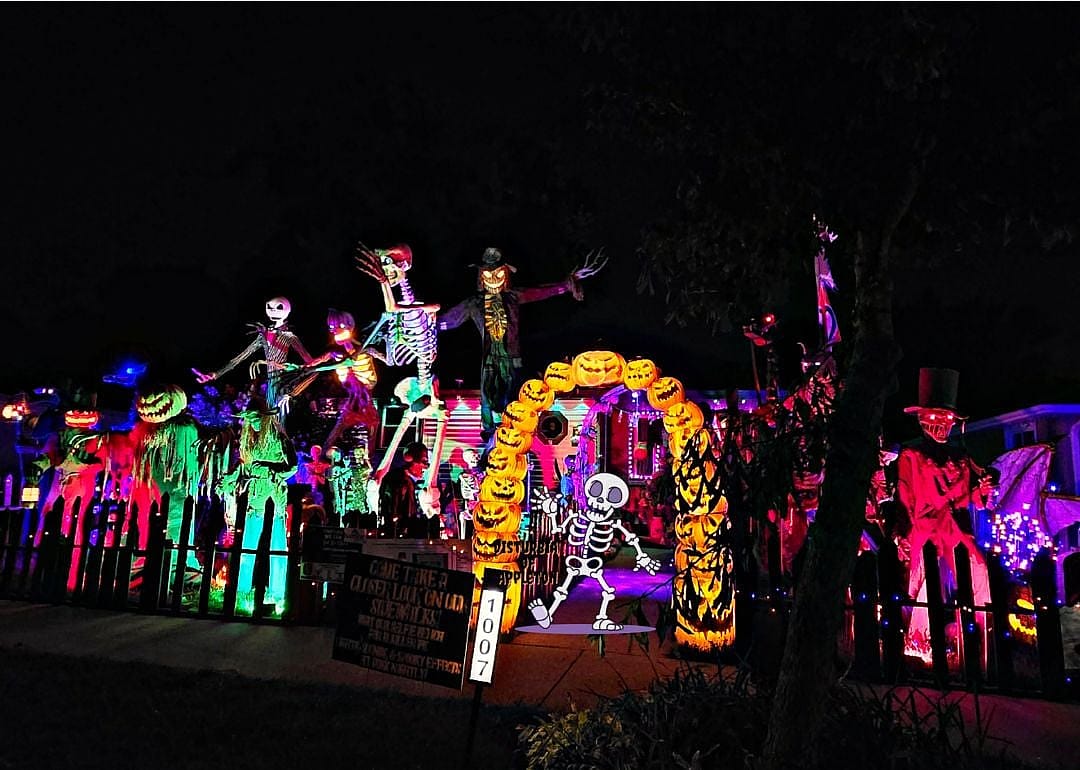 A front yard in Wisconsin decorated with an arch of spooky jack-o’-lanterns and a colorful nighttime display of towering skeletons, sticky spider silk, pumpkins, customized skeletons lit up.