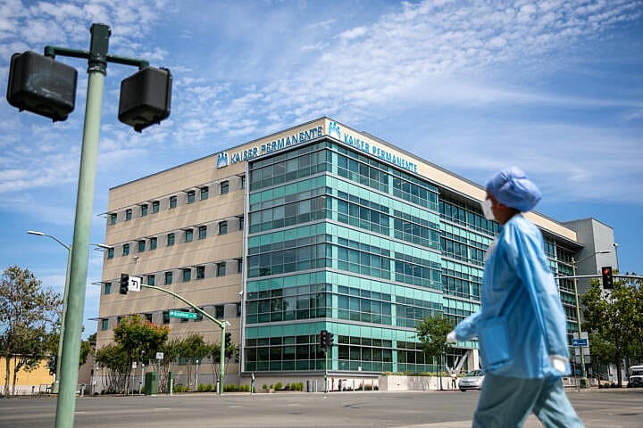 A medical professional in blue scrubs, a surgical mask and hair covering, walks in front of the Kaiser Permanente building in Oakland on Aug. 2, 2022.
