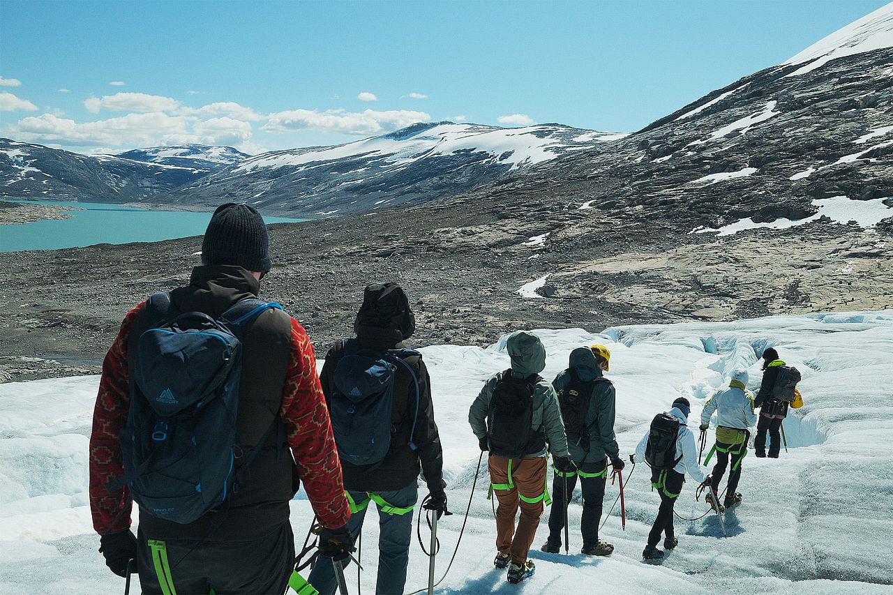 A group of hikers trekking across the blue ice of a glacier in Norway, equipped with safety gear like harnesses and ropes.