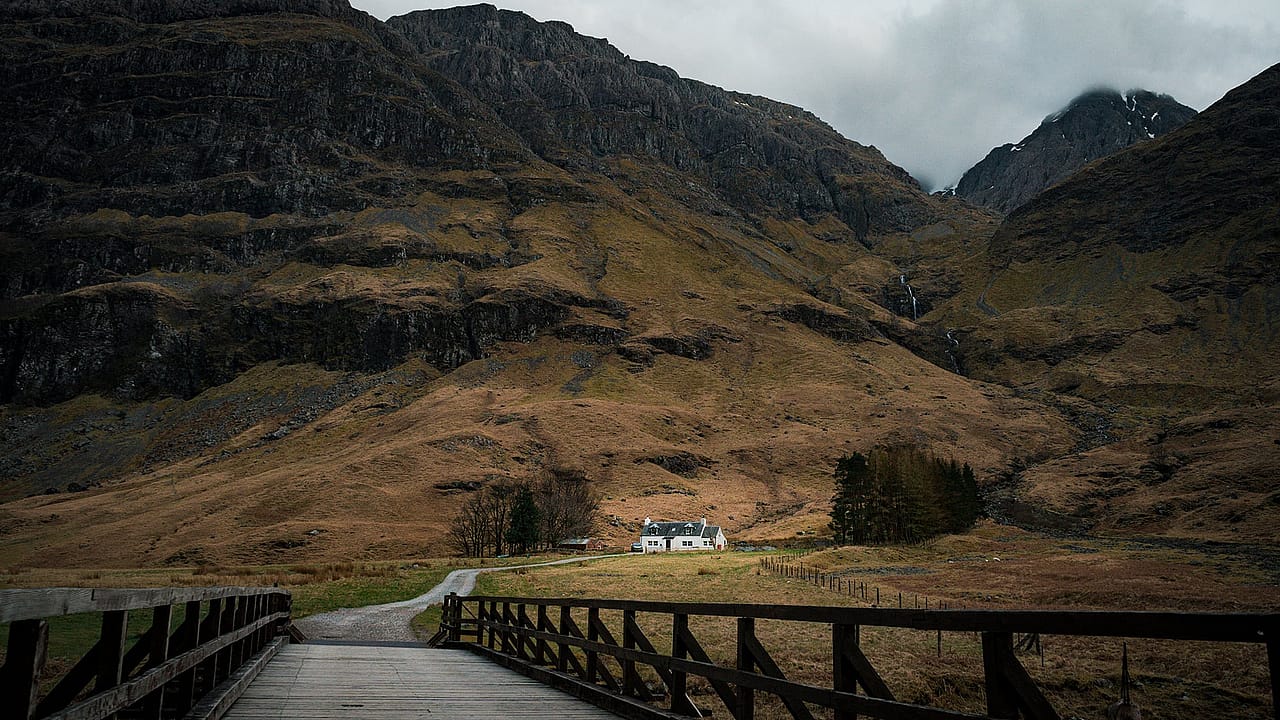 The image shows Achnambeithach Cottage set against the dramatic mountain backdrop near Loch Achtriochtan in Glencoe, Scottish Highlands.