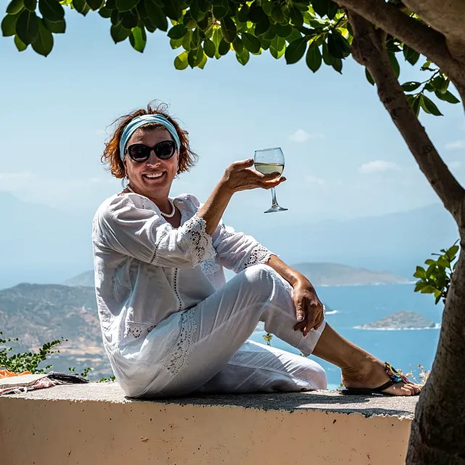 A woman sitting on a stone ledge overlooking a scenic coastal and sea view in the Greek Islands while holding a glass of wine.