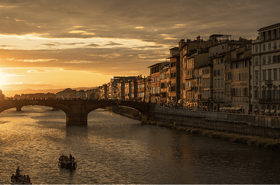 A scenic sunset view over the Arno River in Florence, Italy, looking toward the historic bridges and riverside buildings.