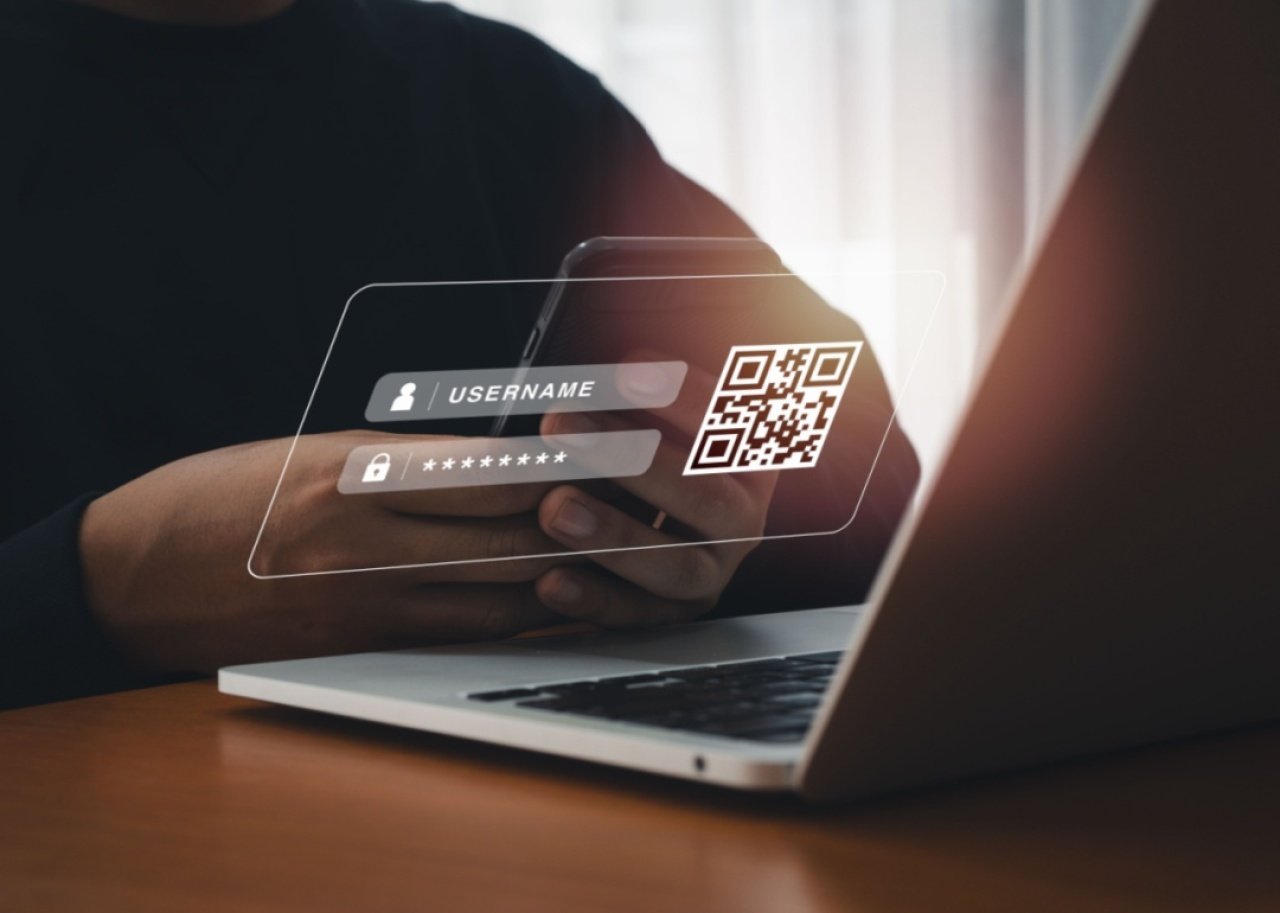 A person holding a smartphone in front of a laptop with an overlaid image of a log in screen and QR code. 