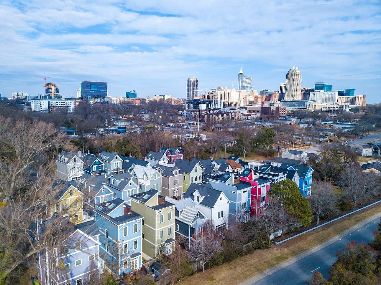 An expansive aerial view of the downtown Raleigh, North Carolina skyline captured from a northern perspective. The photograph showcases the city's prominent skyscrapers, including the PNC Plaza and Wells Fargo Capitol Center, rising against a soft blue sky. In the foreground, a cluster of modern, colorful high-density residential homes provides a stark contrast between neighborhood growth and the metropolitan core. This shot perfectly illustrates the rapid urban development and residential expansion definin