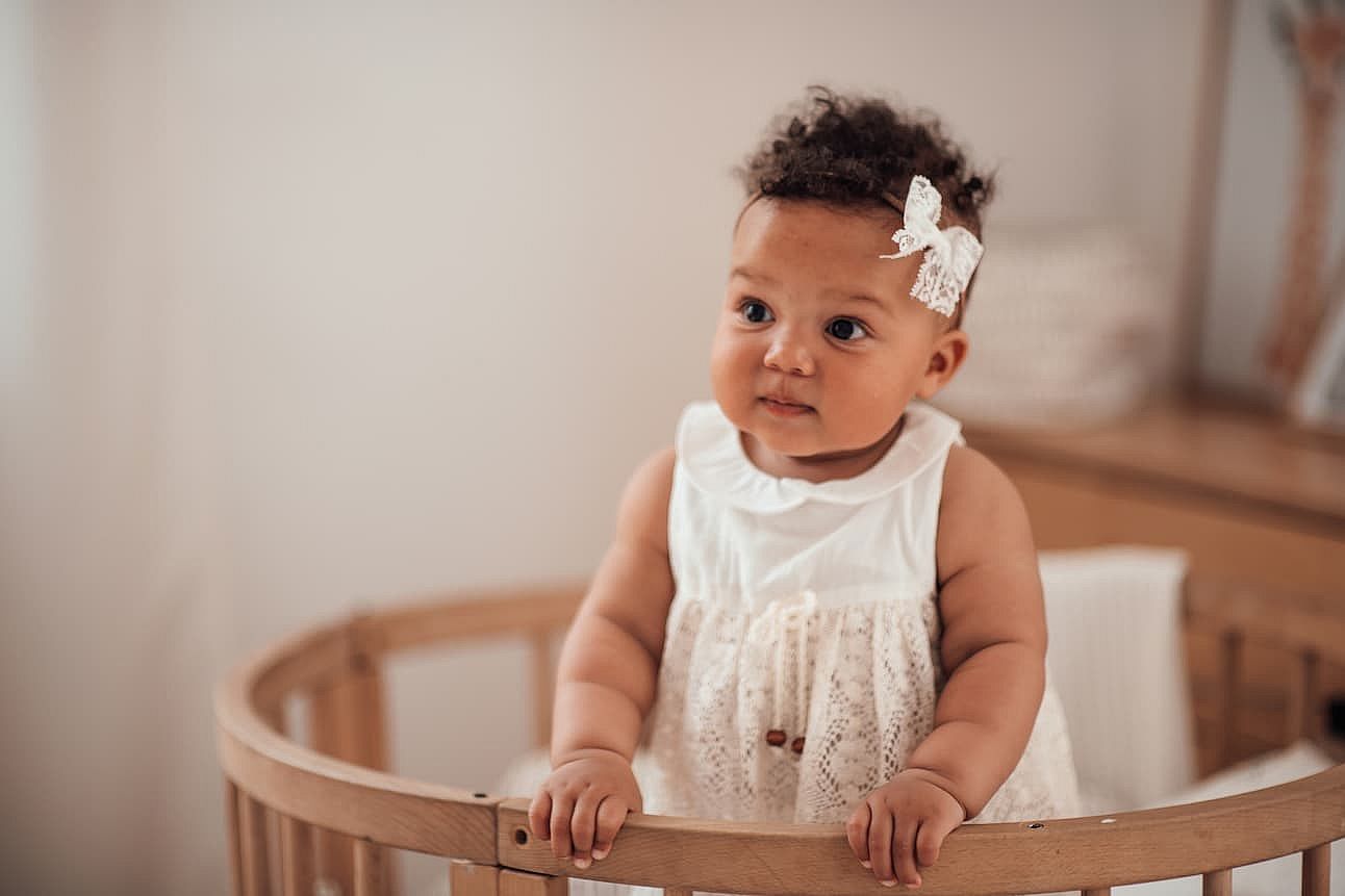 Candid authentic portrait of cute african american diverse female child at home interior of earth colors. Happy swarthy infant baby stands in kids bed in white lace dress and bow headband at bedroom
