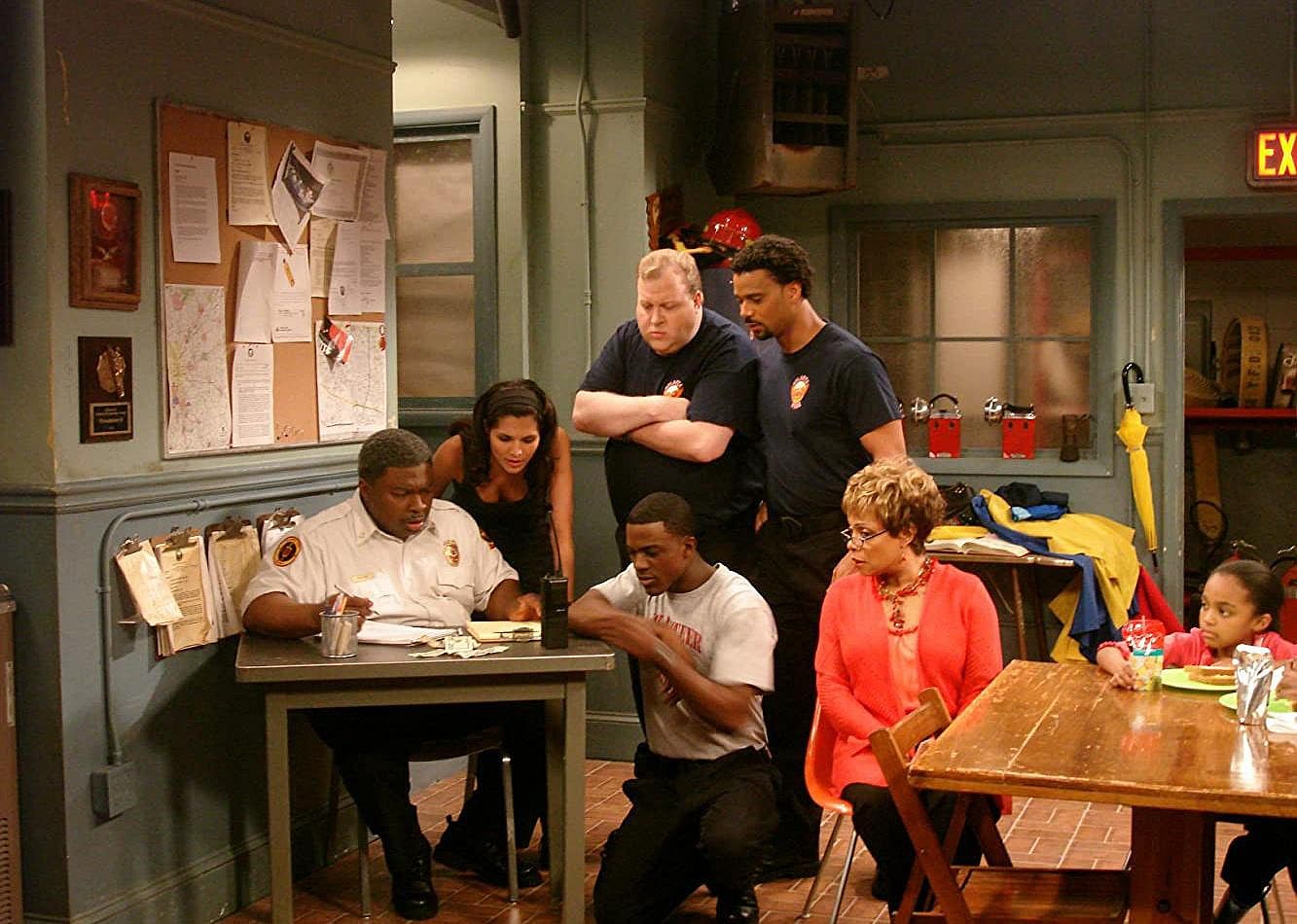 A group of adults and children in a firehouse sit around examining some papers.
