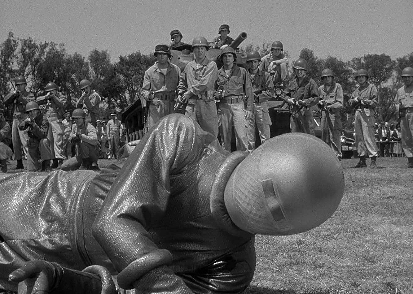 A line of military forces looking at a being in a shiny silver suit on the ground.