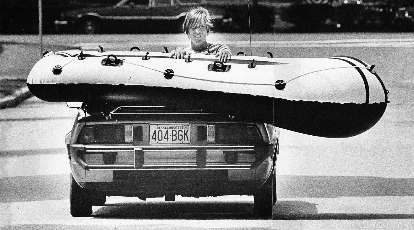 Black-and-white photo of Betsey Cook sitting backward in a car, holding an inflatable boat in place on top of the vehicle as she's driven to the beach in Marblehead, Massachusetts, July 1979.