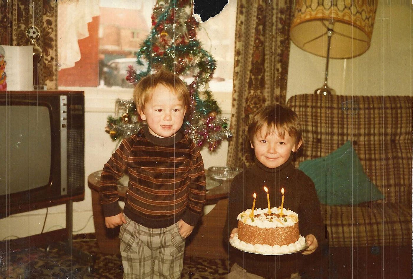 Two boys stand in a brown-toned living room with a Christmas tree behind them, one holding a birthday cake with three lit candles, 1982.