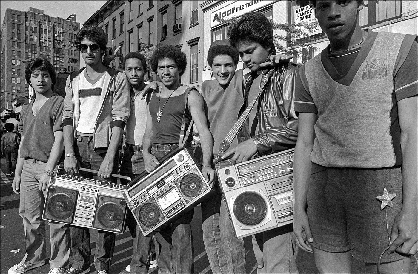 Seven young men pose together on a New York City street, three holding large cassette boomboxes, at the 1982 Ninth Avenue Street Fair.