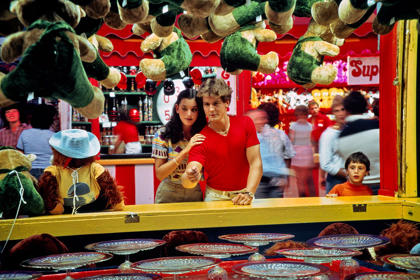 A teenage couple leans over a carnival penny-pitch game surrounded by hanging stuffed animal prizes, while a young boy watches from the side.
