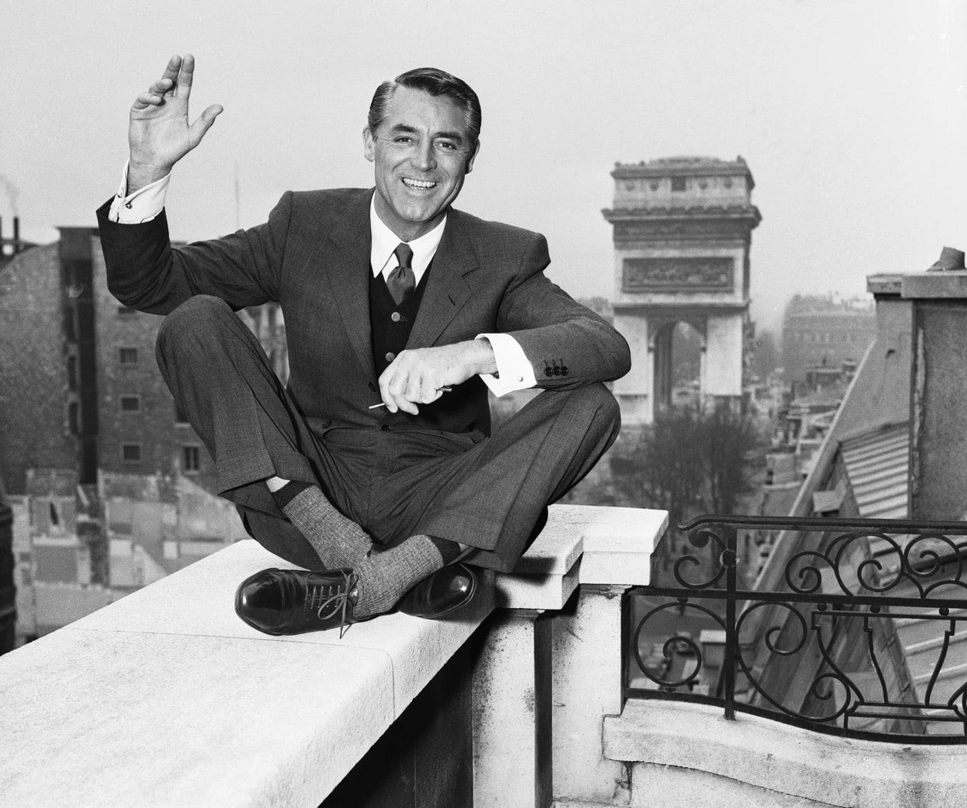 Black and white photo of Cary Grant waving and sitting cross-legged on a balcony in Paris.