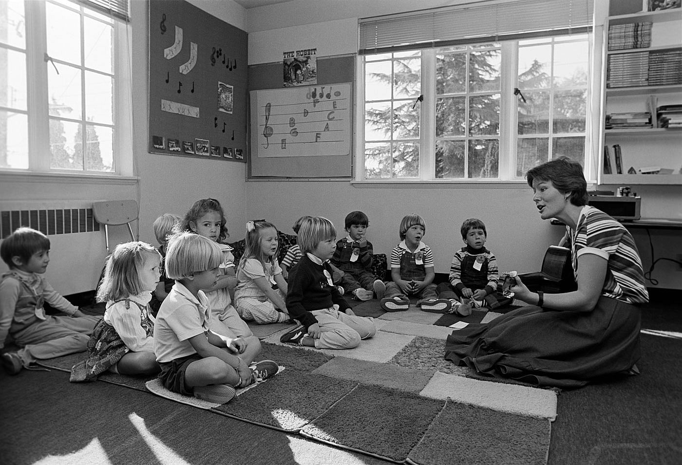 A black and white photo of a teacher playing guitar and singing to a small class of kids.