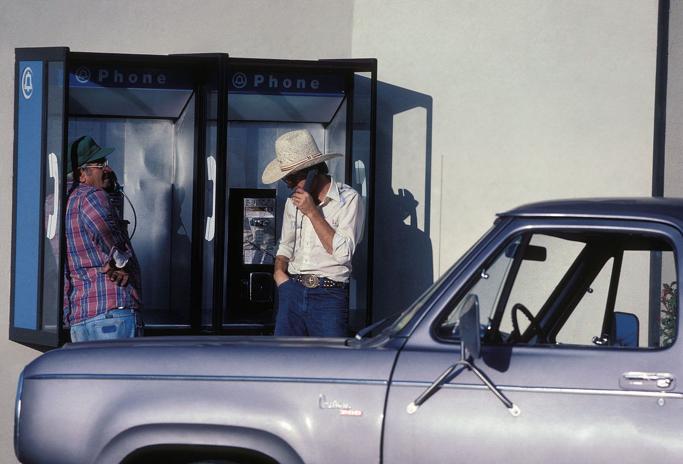 Two men in cowboy hats use side-by-side payphone booths next to a parked pickup truck in Dallas, Texas, November 1982.