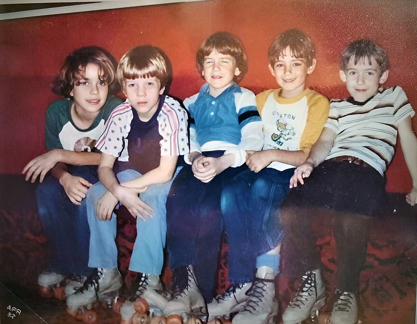 Five young boys wearing roller skates sit closely together on a bench in front of a red wall, 1982.