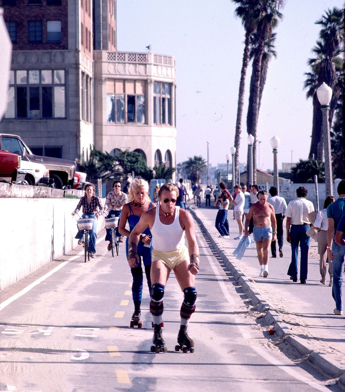 A man and woman roller skate down the Venice Beach boardwalk past cyclists and pedestrians, with palm trees and an oceanfront building behind them, November 1982.