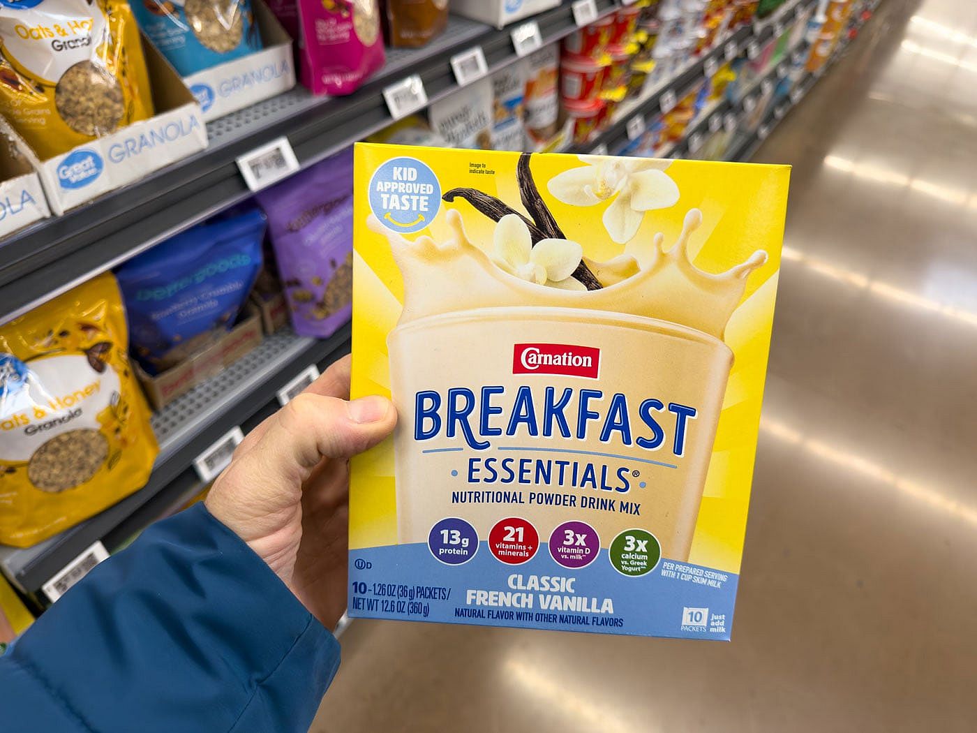 A hand holding a box of Carnation Instant Breakfast Essentials in a grocery store aisle.