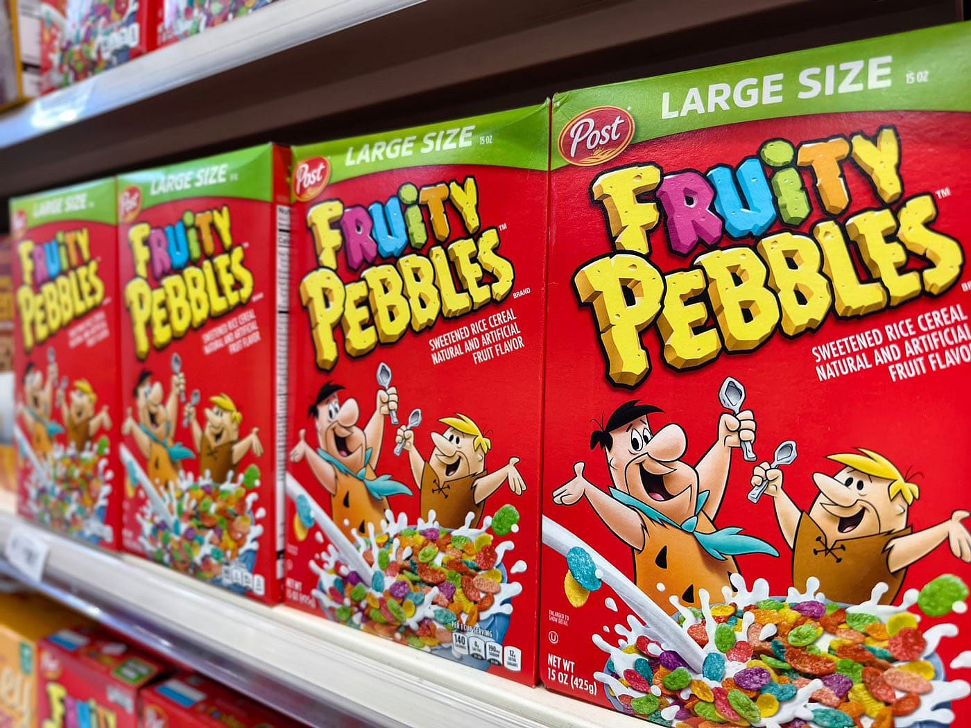 Boxes of Large Size Fruity Pebbles cereal sitting on a shelf at a grocery store.