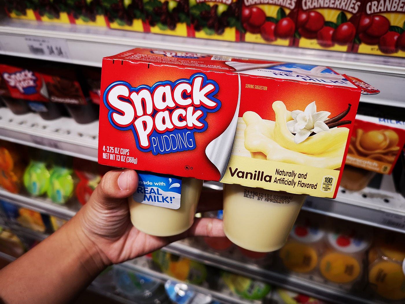 A hand holding a 4-pack of Vanilla Snack Pack puddings in a grocery store aisle.