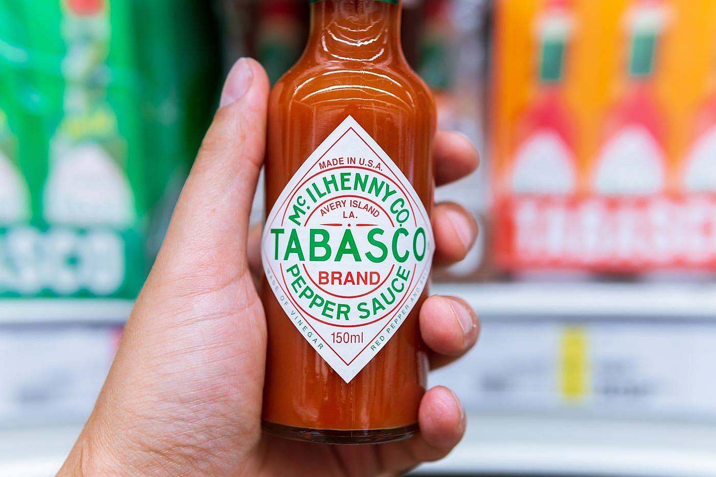 A hand holding a bottle of Tabasco in the hot sauce section of a grocery store.