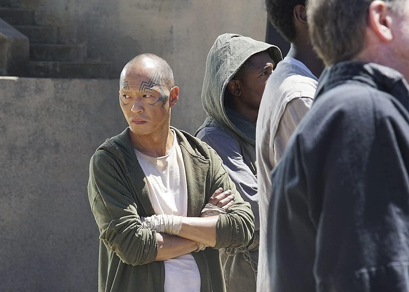 A bald man with tattoos around his eyes and bandages over his knuckles looks back while standing in a group of people looking forward.
