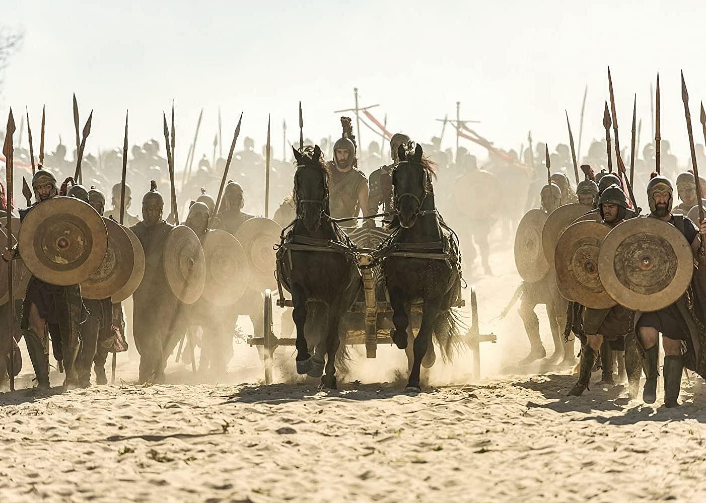Men with shields walk next to two men behind horses.