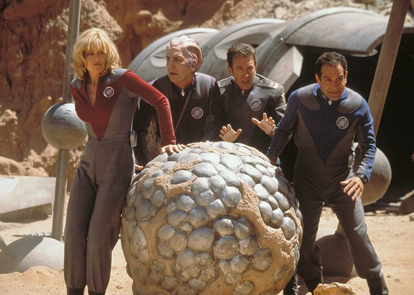 Sigourney Weaver, Alan Rickman, Tim Allen, and Tony Shalhoub, all in space uniforms, stand frightened next to a huge rock.