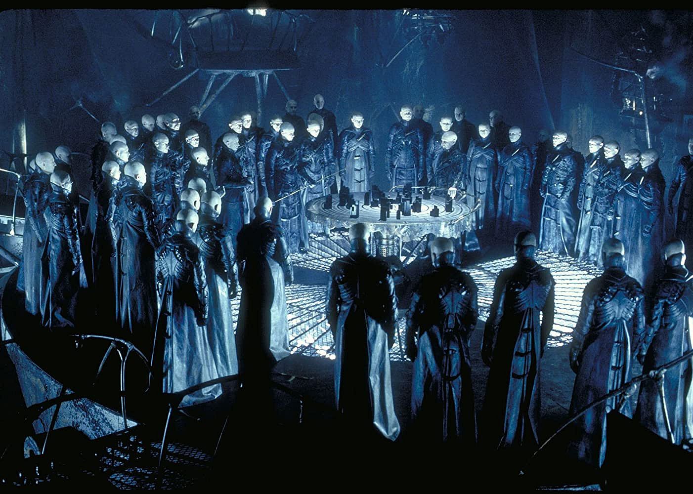 A group of bald beings in dark coats standing in a circle.