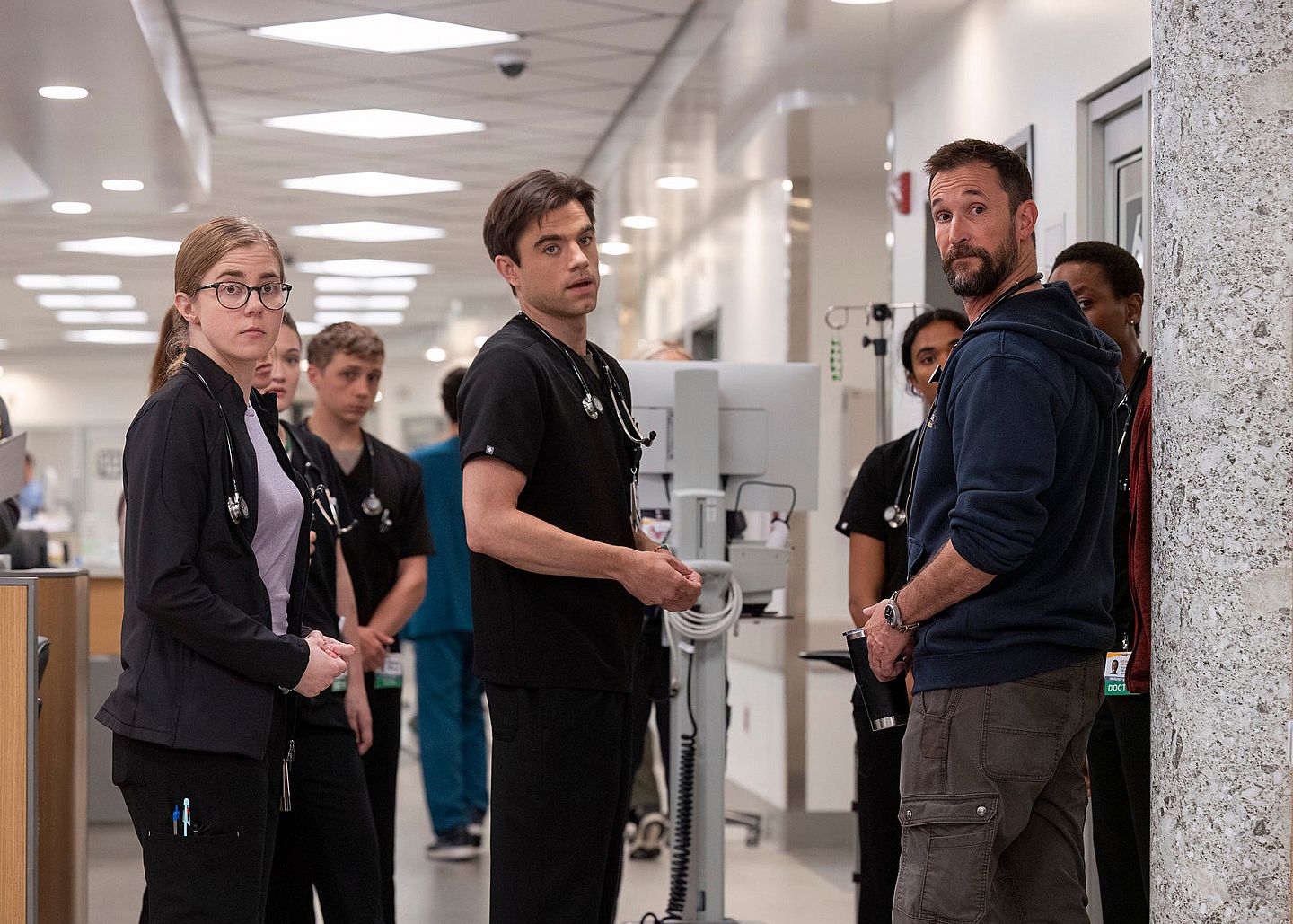 Actors from Season 1 of the TV series “The Pitt” stand in a brightly lit hospital corridor wearing scrubs and ID badges. They appear concerned or surprised, with medical equipment and patient rooms visible behind them.