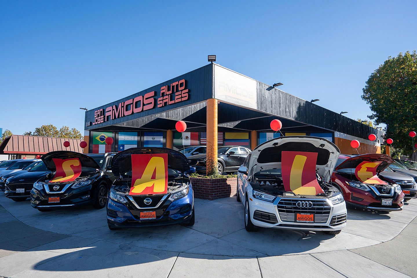 Used car lot in San Jose called Amigos with four cars out front with their hoods up to hold signs that spell out SALE