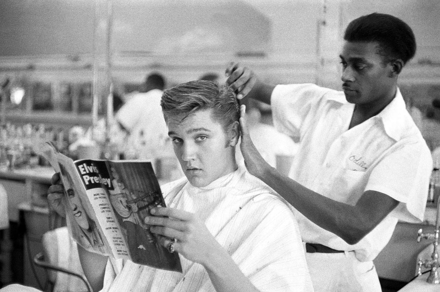 Rock and Roll musician & actor Elvis Presley reads a magazine as barber Odell Elliott cuts his hair in Jim's Barber Shop, Memphis, Tennessee, July 1956.