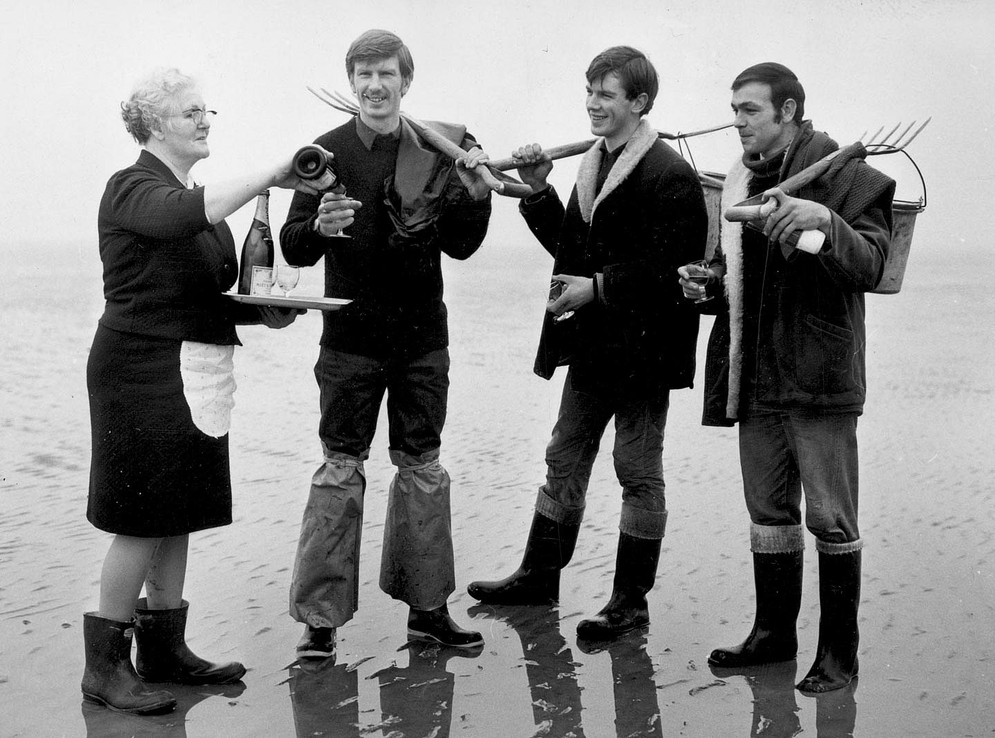 A woman serves champagne to three men in waders who are digging for clams. 