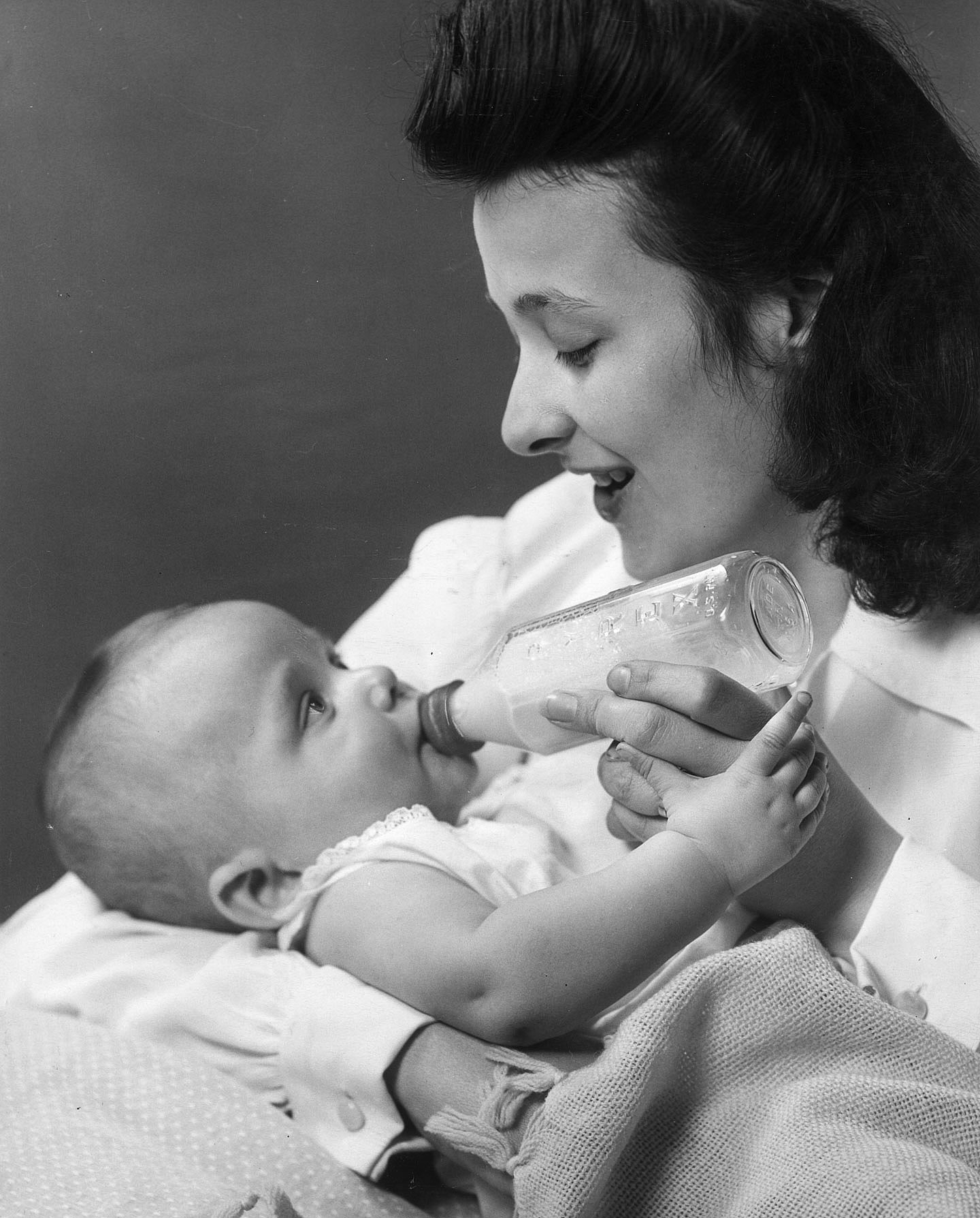 A woman smiles as she feeds holds her newborn baby and feeds her from a bottle of milk, circa 1945.