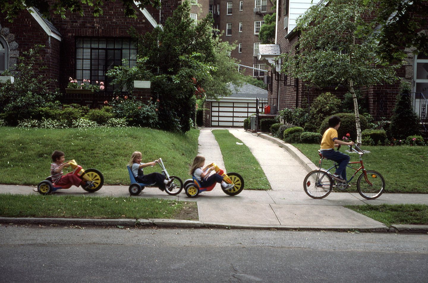 Three young children ride yellow-and-red Big Wheel tricycles in a line along a sidewalk while an older boy rides a bicycle beside them, in front of brick houses on a tree-lined street.