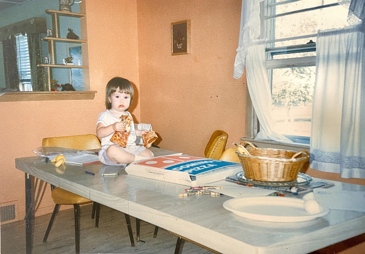 A toddler sits cross-legged on a dining table, holding a slice of pizza in each hand, next to an open Domino's Pizza box. Crayons, a banana, and a wicker basket are scattered around on the table.