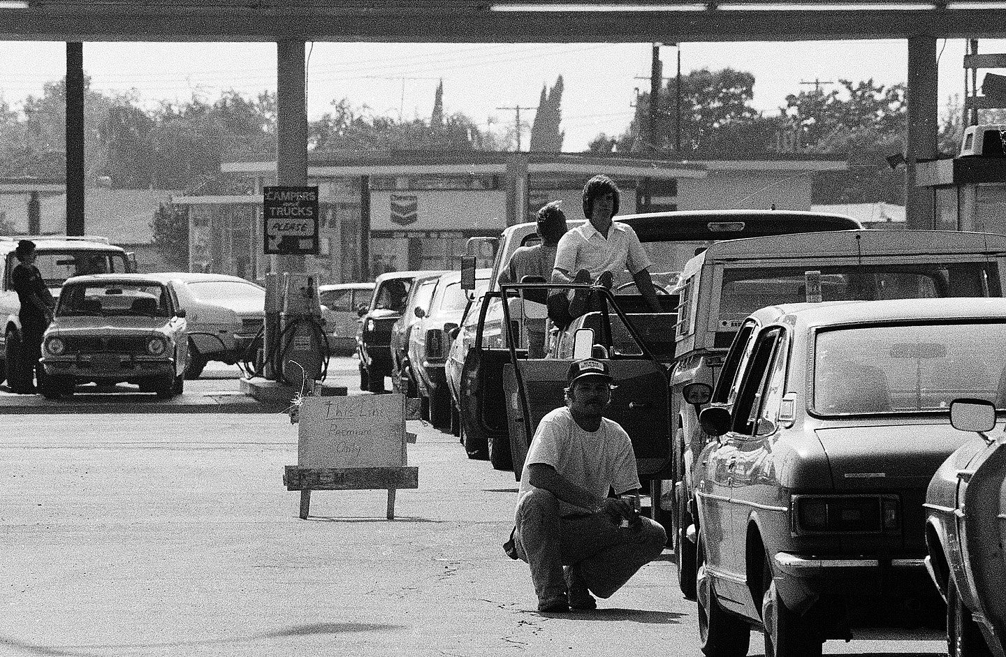 Black-and-white photo of cars lined up bumper-to-bumper at a gas station during the 1979 fuel shortage in Long Beach, California, with a hand-lettered sign reading "This line: Premium only."