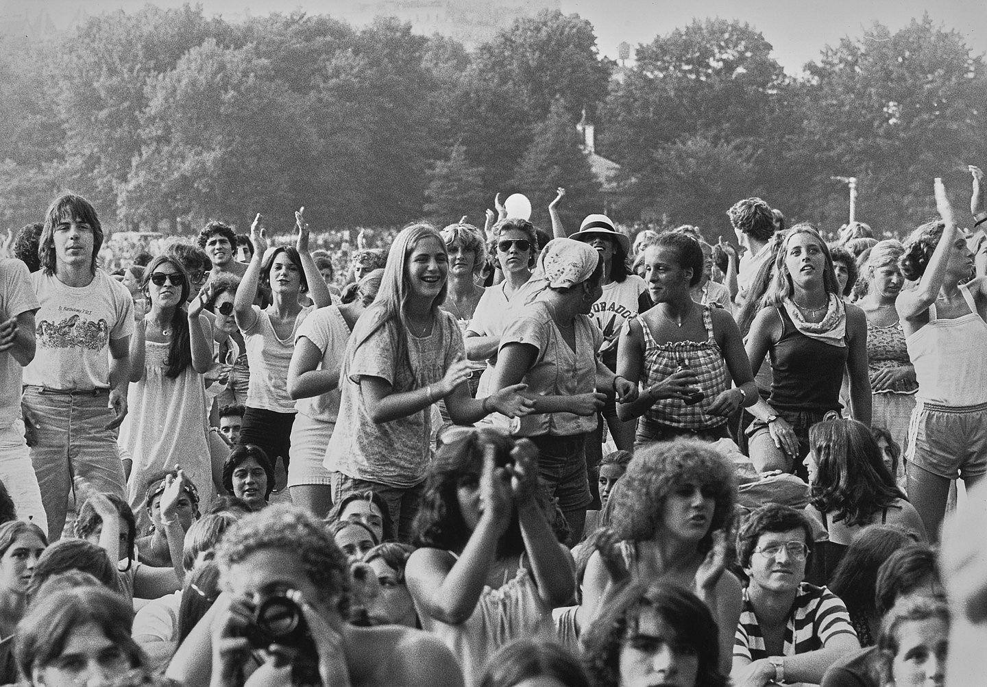 Black-and-white photo of a large crowd dancing, clapping, and taking photos during James Taylor's free concert at Central Park's Sheep Meadow, New York City, July 1979.
